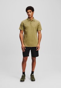 Olive zelené polo tričko s trojitou manžetou, krátkymi rukávmi, logom, v kombinácii s čiernymi šortkami a zelenými športovými topánkami.