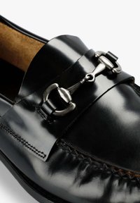 Mocasín de cuero negro con un acabado brillante. Presenta un acento de hebilla metálica y un detallado de costura a lo largo del borde. Forro interior texturizado visible.