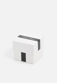 Biały kwadratowy pudełko na prezent z czarną wstążką z napisem "Calvin Klein". Gładkie, matowe wykończenie, prosty design i brak zauważalnych wzorów.