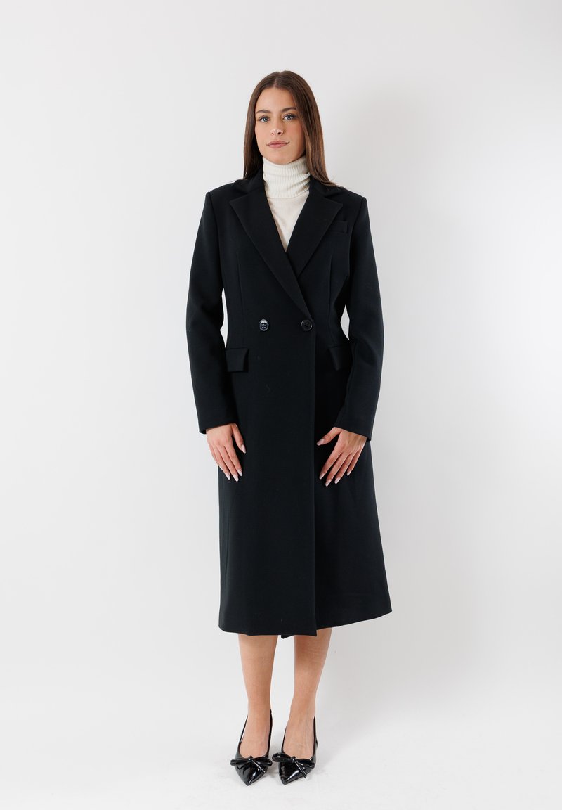 Cappotto nero doppiopetto con rever a punta, tasche laterali e lunghezza fino al polpaccio. Ha una texture liscia ed è abbinato a tacchi a punta neri.