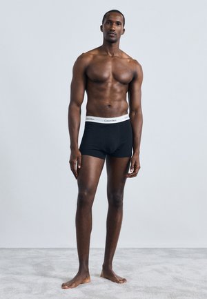 Černé bavlněné boxerky s bílým pasem Calvin Klein. Hladká textura, přiléhavý střih a délka do poloviny stehna. Model stojí bos.