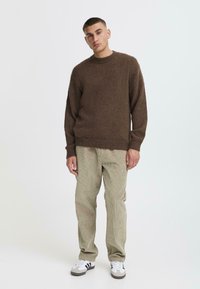 Brūns adīts džemperis ar apaļu kaklu, brīvs piegriezums. Apvienots ar gaiši zaļām kordpantaloniem un baltām sporta kedām ar melnām svītrām.