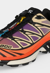 Chaussure de trail avec panneaux violets, orange et beige, système de laçage rapide noir et semelle texturée noire et blanche.