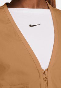 Béžová zipová vesta s texturovaným povrchem přes bílý tričko s kulatým výstřihem, na kterém je černé logo Nike. Důraz na jednoduchost a funkčnost.