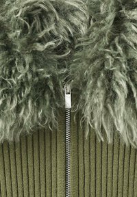 Chaqueta de tela texturizada verde con un collar esponjoso, un zipper plateado en el frente y detalles acanalados en el cuerpo principal.