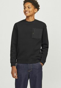 Svart sweatshirt med rund halsringning, långa ärmar och en bröstficka framtill, tillverkad av ett strukturerat tyg. Buren med mörkblå jeans.