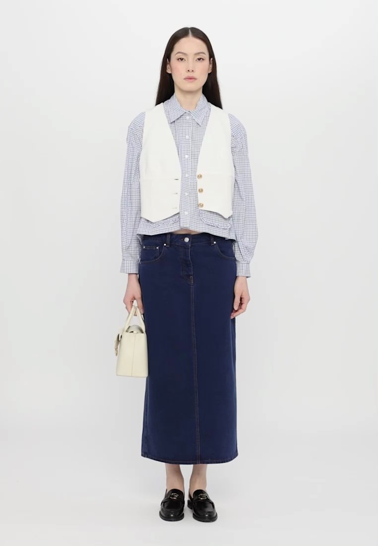 Gilet blanc sur une chemise à carreaux bleue, associé à une longue jupe en denim foncé. Les accessoires comprennent un petit sac blanc et des chaussures noires.