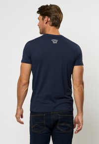 Marineblå kortermet T-skjorte, laget av bomull, rund hals, med hvit "AUTHENTIC" logo på øvre rygg. Brukt med mørke jeans.