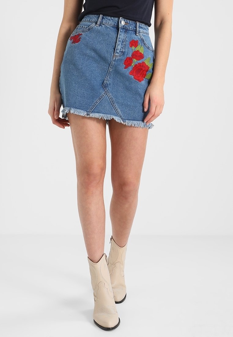 Femme portant une jupe en jean bleue avec une broderie de rose rouge et un ourlet effiloché, associée à des bottines beiges.