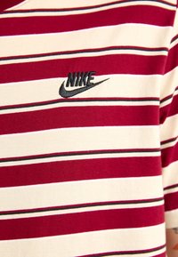 Punainen ja valkoinen raidoitettu t-paita, joka on valmistettu puuvillasta, sisältää ohuita mustia raitoja, ribattu kaulus ja musta Nike-logo rinnassa.