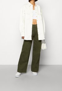Persona vestida con una camisa de botón blanca oversize sobre un crop top blanco, pantalones anchos oliva, zapatillas blancas, sosteniendo un pequeño bolso blanco.
