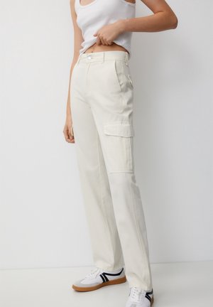 Nainen, jolla on päällään luonnonvalkoiset cargo-housut, valkoinen toppi ja valkoiset mustaraidalliset tennarit, seisoo yksiväristä vaaleaa taustaa vasten.