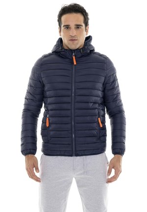 Un homme porte une veste matelassée à capuche bleu marine, avec des tirettes de fermeture éclair orange, et un pantalon de jogging gris clair sur fond blanc.