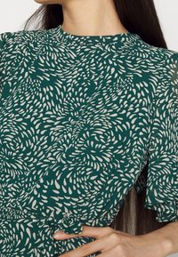 Vihreä mekko, jossa on valkoinen pyörivä kuvio, lyhyet röyhelöhihat, pyöreä pääntie ja vyö, valmistettu kevyestä kankaasta.