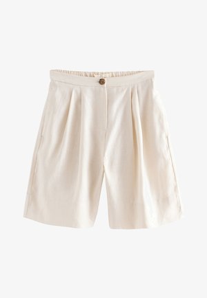 Donna che indossa pantaloncini larghi in lino beige a vita alta con pieghe, un bottone marrone e un top beige corto, mano in tasca, braccialetti dorati al polso.