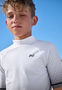 Vit rashguard med kort ärm och polokrage, med ett gradientmönster av svarta prickar och en liten svart logotyp på bröstet.