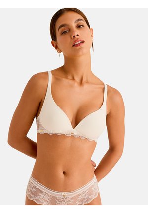 Femme portant un ensemble assorti de soutien-gorge et culotte en dentelle couleur crème, posant les mains derrière le dos sur un fond uni.