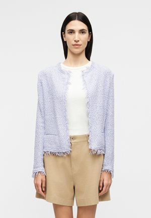 Egyenes sötét hajú nő világos levendulás, texturált, nyitott elülső részű dzsekit, fehér felsőt és bézs rövidnadrágot visel, egyszínű háttér előtt állva.