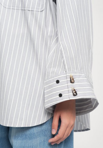 Gray striped shirt with white lines, featuring a relaxed fit, black button accents, and unique cuff detailing.
