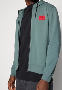 Muž nosící rozepnutou tyrkysovou mikinu s kapucí a červenou nášivkou HUGO přes černé tričko a tmavě šedé kalhoty, stojící před jednobarevným pozadím.