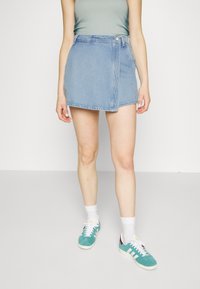 Falda mini de mezclilla, azul claro, estilo envolvente con cierre de botón, combinada con una blusa verde ajustada y zapatillas de deporte en color azul verdoso. Textura de tela plana y suave.