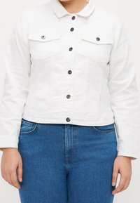 Person som bär en vit knäppt jacka med framfickor och blå denimjeans, står med armarna avslappnat vid sidorna.