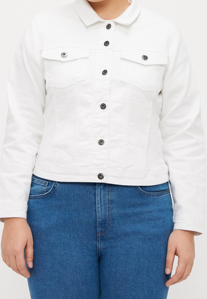 Person som bär en vit knäppt jacka med framfickor och blå denimjeans, står med armarna avslappnat vid sidorna.