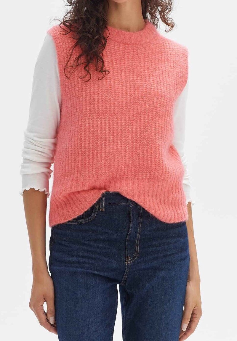 Femme portant un pull sans manches en tricot rose par-dessus une chemise blanche à manches longues et un jean bleu foncé, debout devant un fond uni.