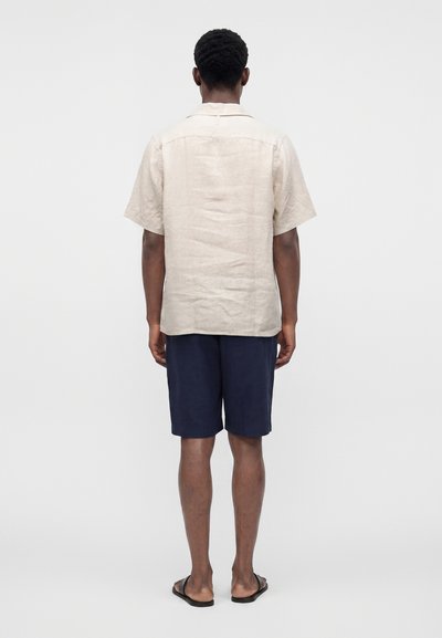 Homme debout de dos, portant une chemise beige à manches courtes, un short bleu marine et des tongs noires sur un fond uni.