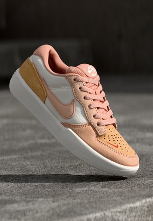 Baskets Nike rose clair et beige avec semelle et lacets blancs, languette marquée, et zone perforée à l'avant sur sol en béton.
