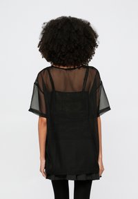 Schwarzes durchsichtiges Oberteil mit kurzen Ärmeln, lockerem Schnitt, Lagen-Design und filigranen Details auf der Rückseite, kombiniert mit einem schwarzen plissierten Rock.