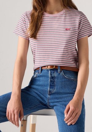 Femme portant un t-shirt Levi's rose et blanc à rayures, rentré dans un jean bleu taille haute avec une ceinture marron, assise sur un tabouret en bois.