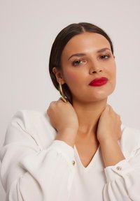 Biała bluzka z bufkami i guzikami, zestawiona ze złotą kolczykiem w kształcie koła. Skóra wygląda gładko, z wyrazistym czerwonym kolorem ust.
