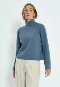 Gaiši zils augstais kakls džemperis no mīksta auduma, ar gar�ām piedurknēm un īsu dizainu, apvienots ar bežām augstām biksēm.