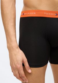 Μαύρα βαμβακερά ανδρικά εσώρουχα boxer με πορτοκαλί ελαστική μέση που φέρει το λογότυπο "TOMMY HILFIGER" σε λευκό. Λείο υφή και εφαρμοστή σχεδίαση.