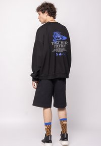 Black oversized sweatshirt with blue graphic text on the back, paired with black shorts and patterned orange and blue socks.