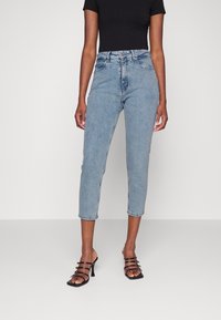 Mujer vistiendo unos jeans de tiro alto cortos de color azul claro, combinados con una blusa negra de manga corta y sandalias de tacón alto negras con tiras.
