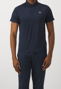 Mężczyzna noszący granatową koszulę polo z krótkim rękawem i małym zielonym logo krokodyla na lewej piersi oraz dopasowane granatowe spodnie.