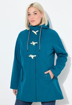 Woman wearing a teal hooded jacket with wooden toggle buttons and white drawstrings, standing with one hand in pocket against white background.