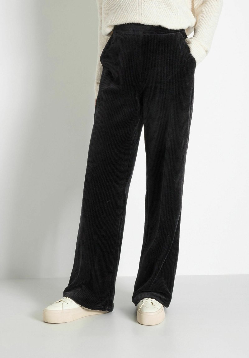 Pantalons en velours côtelé noir à jambes larges avec texture à côtes verticales, dotés de poches avant et d'une taille haute, associés à des baskets à plateforme crème.