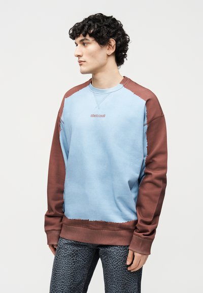 Νέος άνδρας με σγουρά μαλλιά, που φοράει μπλε και καφέ φούτερ Roberto Cavalli και παντελόνια με λεοπάρ σχέδιο, στραμμένος αριστερά πάνω σε απλό φόντο.