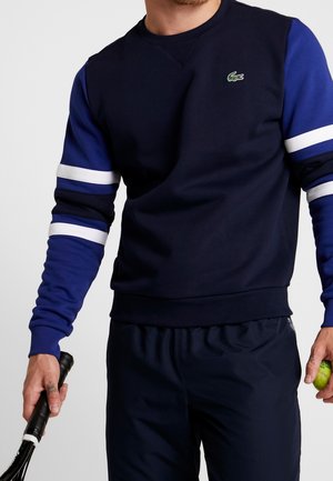 Ein Mann trägt einen marineblauen und blauen Sweatshirt mit weißen Streifen, hält in einer Hand einen Tennisschläger und in der anderen einen Tennisball.