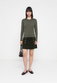 Un haut à manches longues olive associé à une jupe en velours côtelé vert foncé avec des boutons en argent. Les accessoires comprennent un petit sac à main noir et des ballerines en vernis.