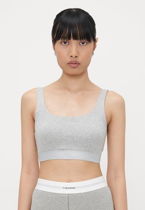 Grey ribbed bralette with wide straps and a logo band. The fabric appears soft with a subtle texture. Fitted design, minimalist style.