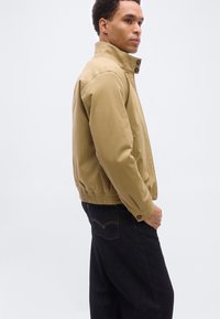 Άνδρας που φοράει μπεζ ζακέτα με φερμουάρ και ψηλό γιακά, καθώς και μαύρο τζιν, στεκόμενος προφίλ μπροστά σε απλό λευκό φόντο.