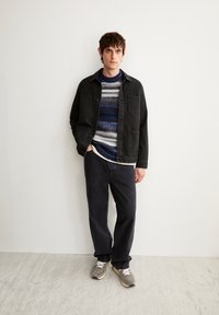 Schwarze Jeansjacke über einem mehrfarbigen, gestreiften Strickpullover; dunkle Hose und graue Sneaker. Minimalistisches Design, lässige Ausstrahlung.