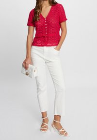 Haut en dentelle rouge à manches courtes, décolleté en V et fermeture à boutons ; jean blanc court et sac à main matelassé blanc avec des accessoires en or.