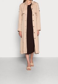 Trench coat beige con grandi bottoni, spalline e una vestibilità morbida, indossato sopra un vestito a coste marrone scuro e sandali piatti intrecciati.