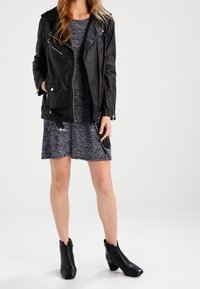 Schwarze Leder-Bikerjacke mit silbernen Reißverschlüssen, über einem strukturierten grauen Kleid getragen. Das Model trägt schwarze Ankle-Boots mit spitzer Zehenpartie.