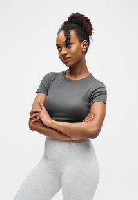 Jeune femme aux cheveux bouclés attachés en un chignon haut, portant un t-shirt gris ajusté et des leggings gris clair, debout les bras croisés et regardant sur le côté.
